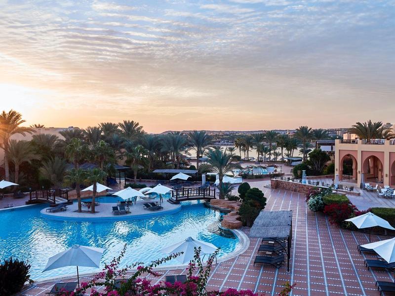 Luxury resort with large pools, palm trees, sun umbrellas, and sunset sky.