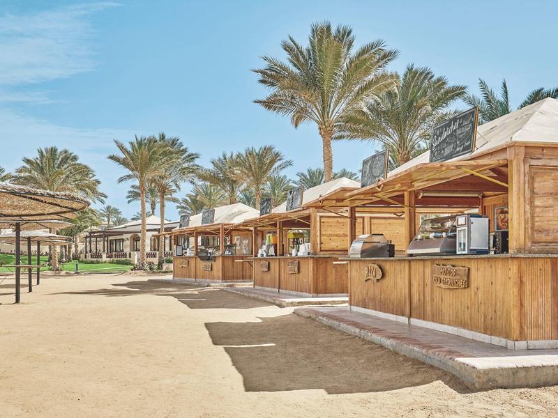 Row of wooden huts with palm roofs on sandy beach under blue sky.