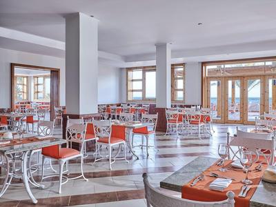 Hell eingerichteter Speisesaal mit weißen Tischen, orangen Stühlen und großen Fenstern mit Blick nach draußen.