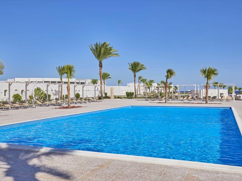 Großer blauer Pool mit Palmen und weißen Gebäuden unter klarem Himmel.