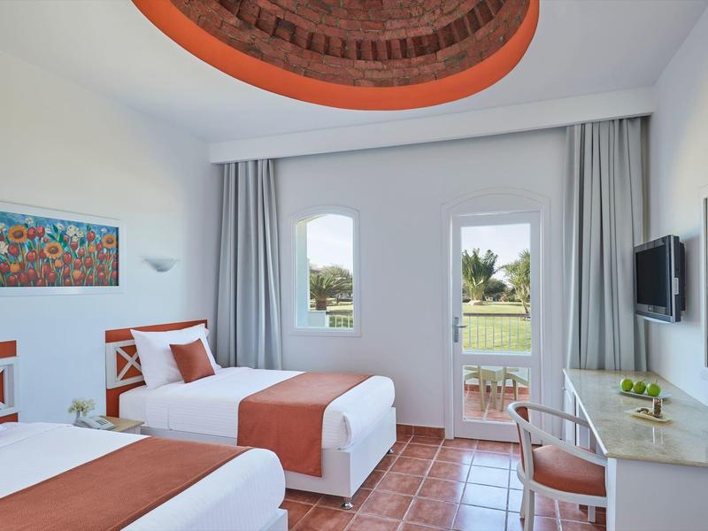 Hell eingerichtetes Doppelzimmer mit zwei Betten, oranger Bettwäsche und Zugang zu Balkon mit Gartenblick.