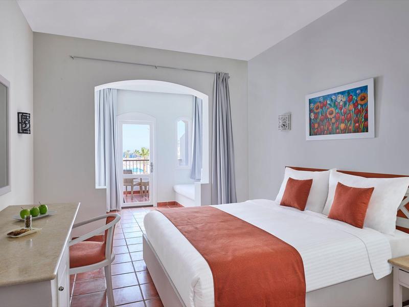 Helles Schlafzimmer mit Doppelbett, oranger Bettdecke, zwei Stühlen und Blick auf Balkon und Meer.