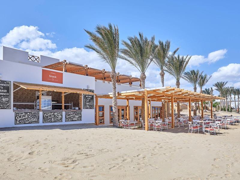 Strandrestaurant mit Holzdach, Palmen und Sand unter blauem Himmel und weißen Wolken.
