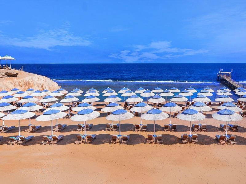 Strand mit vielen weißen Sonnenschirmen und Liegen vor blauem Meer und klarem Himmel.