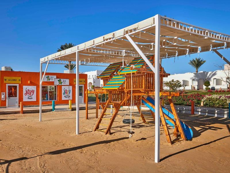 Kinderspielplatz mit Holzgerüst, Rutsche, Sandboden und weißem Sonnenschutzdach bei klarem Himmel.