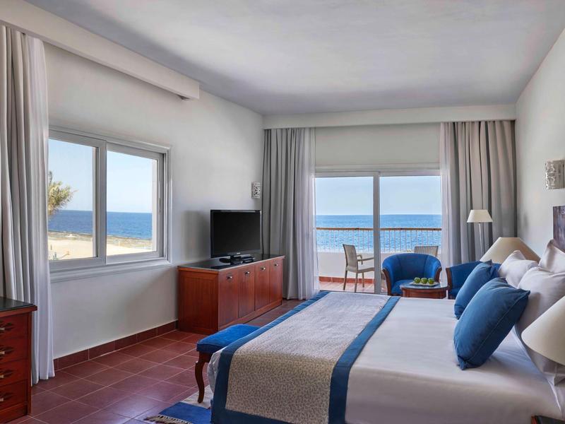 Helles Schlafzimmer mit großem Fenster, Meerblick, Doppelbett und Holzmöbeln.