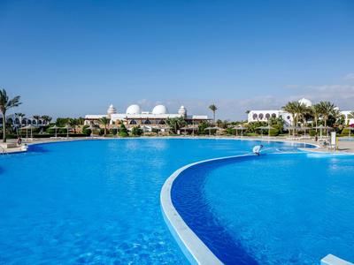 Großer blauer Pool mit klarer Wasserfläche, Palmen und weißem Gebäude im Hintergrund unter blauem Himmel.