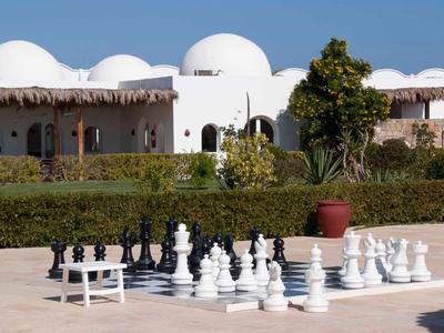 Großes Schachspiel im Freien vor weißem, kuppelförmigem Hotelgebäude und grünen Büschen.