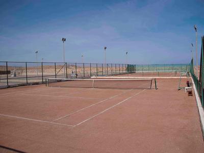 Freiluft-Tennisplatz mit Sandboden und Meerblick bei wolkenlosem Himmel.