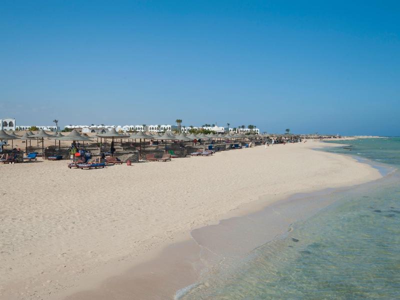 Une large plage de sable avec de l'eau claire et des chaises longues en arrière-plan sous un ciel bleu.