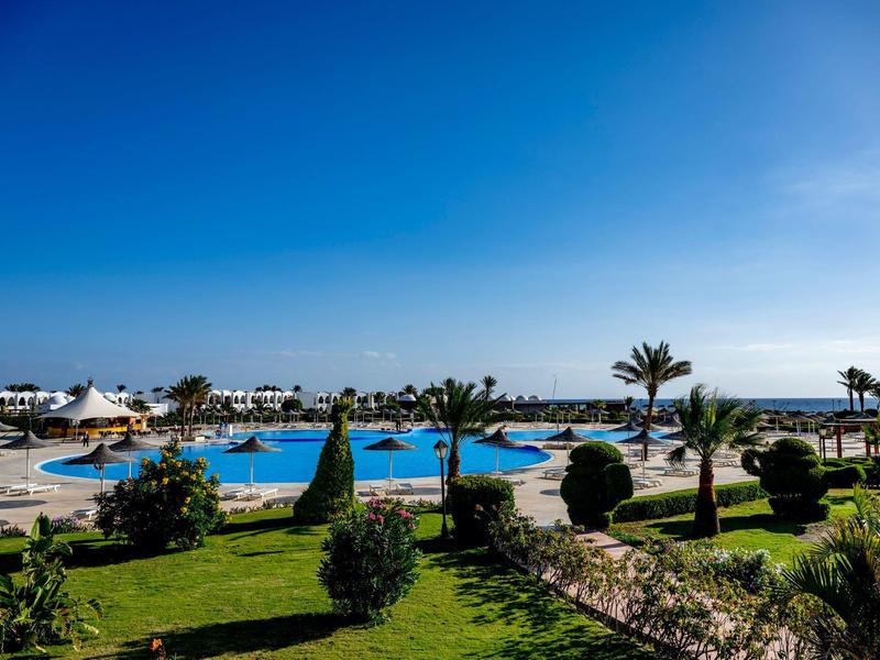 Grande piscine d'hôtel avec palmiers, chaises longues et vue sur la mer sous un ciel dégagé.
