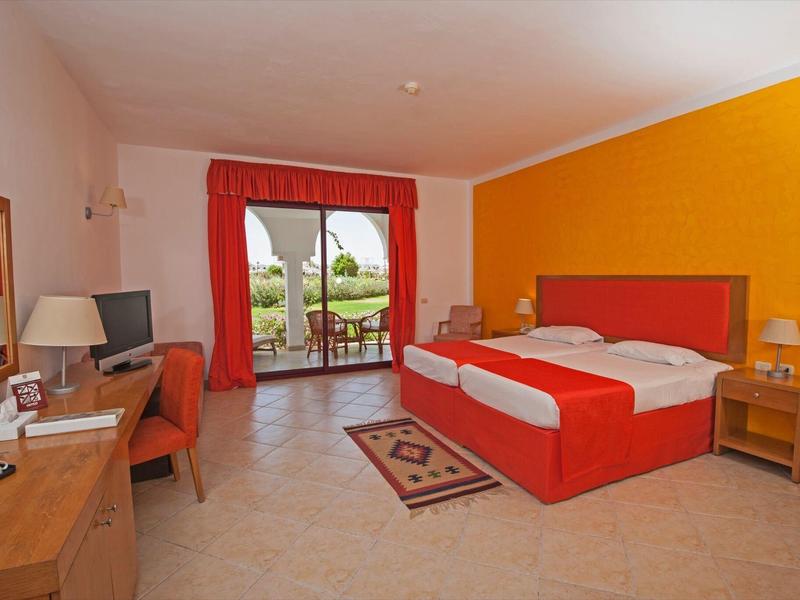 Chambre d'hôtel avec lit rouge, mur orange et accès à une terrasse avec vue sur la verdure.