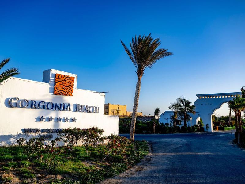 Panneau d'entrée et chemin menant à l'hôtel Gorgonia Beach avec des palmiers et un ciel bleu clair.
