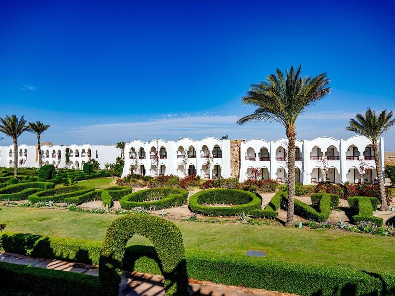 Bâtiment d'hôtel blanc avec jardin bien entretenu et palmiers sous un ciel bleu clair.