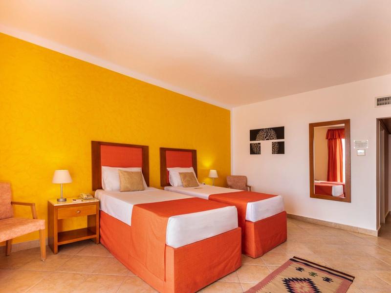 Chambre d'hôtel avec deux lits simples, mur jaune et mobilier simple.