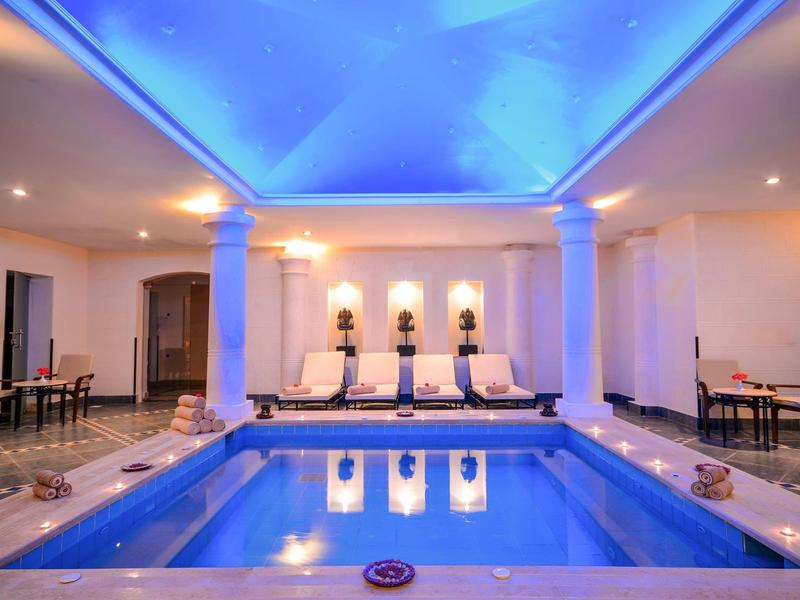 Indoor pool with blue-lit water, lounge chairs, and tables in a hotel setting.