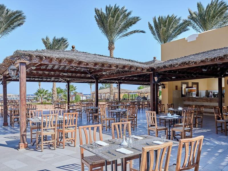 Outdoor restaurant area with wooden tables, chairs, and palm trees in the background.