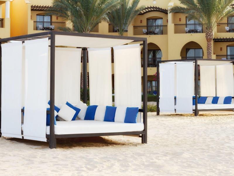 White beach beds with blue pillows on sand in front of hotel with palm trees and balconies.