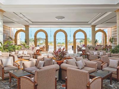 Heller, offener Lounge-Bereich mit beige Sesseln, Pflanzen und großen Bogenfenstern mit Meerblick.