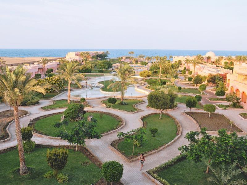 Grüner, gepflegter Garten mit Palmen, Wegen und Brunnen vor Meer und klar blauem Himmel.
