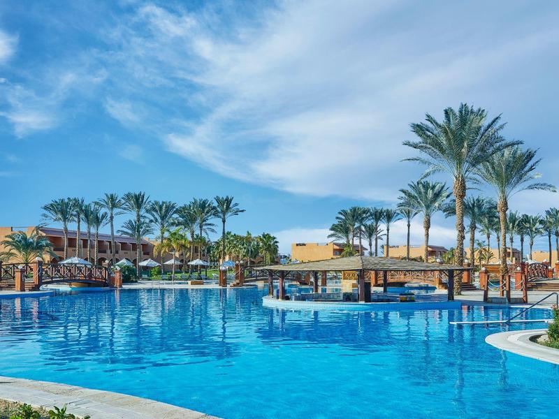 Großer blauer Pool mit Palmen und Sonnenschirmen unter blauem Himmel in tropischer Umgebung.
