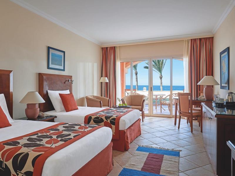 Helles Hotelzimmer mit zwei Betten, bunten Decken, Tisch, Stühlen und Meerblick durch große Fenster.