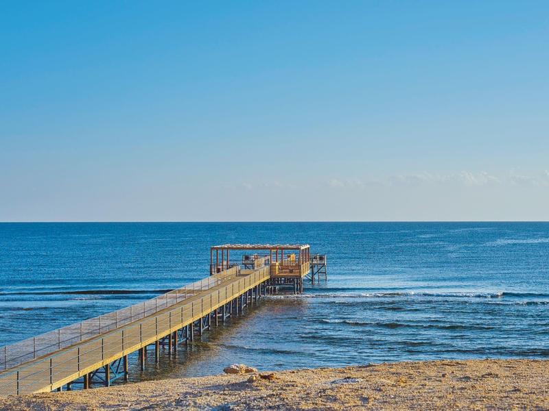 Langer Holzsteg führt über Sandstrand hinaus ins ruhige blaue Meer unter klarem Himmel.