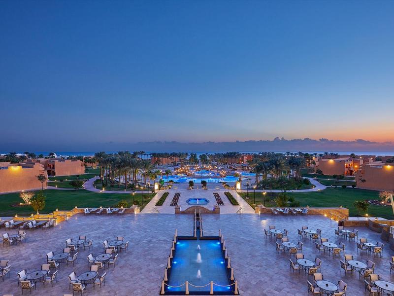 Große Terrasse mit weißen Tischen und Stühlen, beleuchteten Brunnen und Blick auf dahinter liegendes Meer.