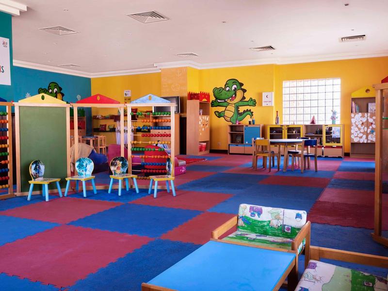 Bunter Kindergartenraum mit weichem Teppich, kleinen Tischen, Spielzeug und Sitzbänken.