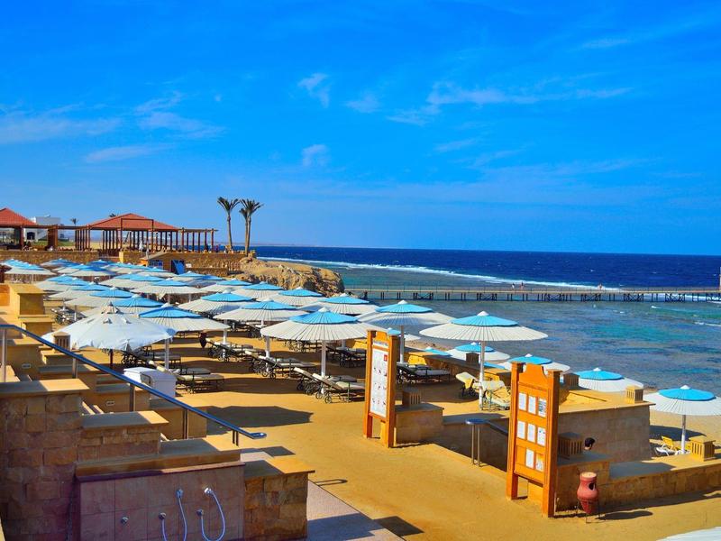 Strand mit mehreren blauen Sonnenschirmen, Liegen und Palmen am klaren blauen Meer unter blauem Himmel.