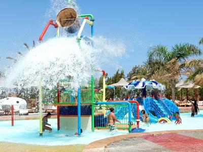 Wasserpark mit Rutschen, Palmen und spreizenden Wasserfontänen unter blauem Himmel.