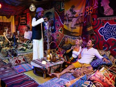 Buntes Zimmer mit Teppichen, Sitzkissen, mehreren Menschen und traditioneller Dekoration an den Wänden.