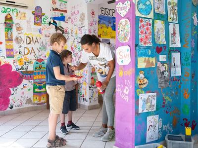 Kinder und Lehrerin besprechen Kunstwerke in einem hellen, farbenfroh bemalten Klassenraum.