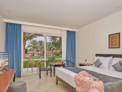 Habitación de hotel luminosa con cama doble, cortinas azules y vista al jardín con palmeras.
