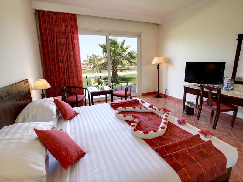 Bright hotel room with a large bed, red accents, and a window showing palm trees outside.