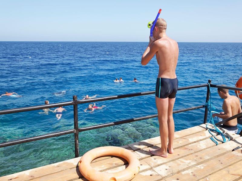 Man with snorkel standing on wooden pier beside clear blue sea with swimmers and life rings.