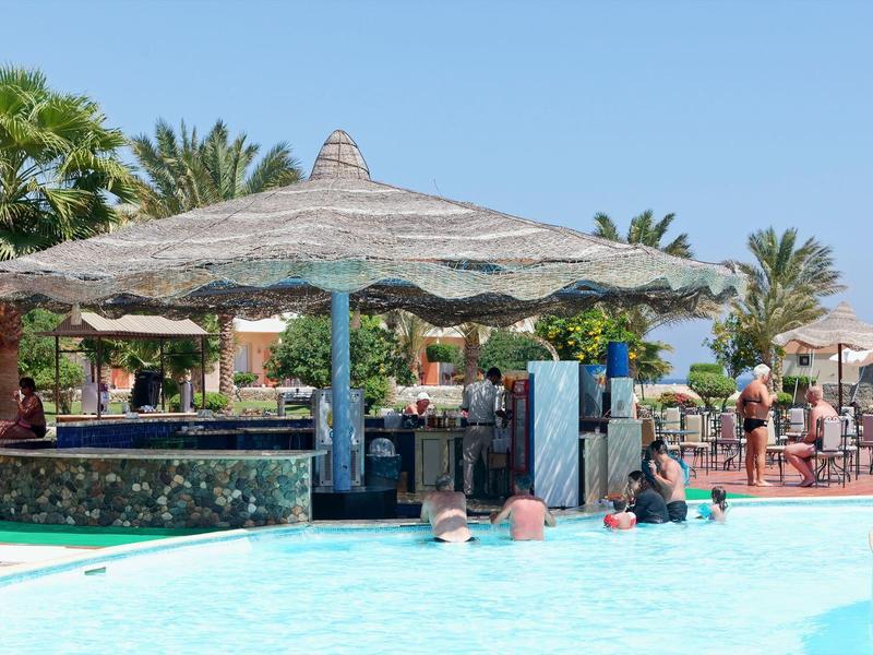 Pool bar with straw roof and people enjoying drinks and swimming under clear blue sky.