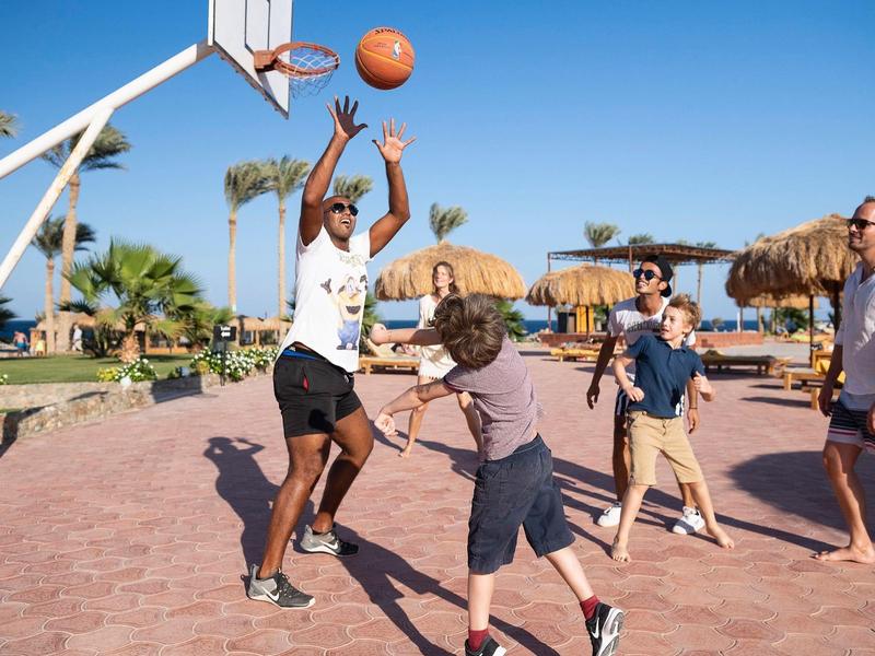 People playing basketball outdoors on a sunny day with palm trees and thatched umbrellas in the background.