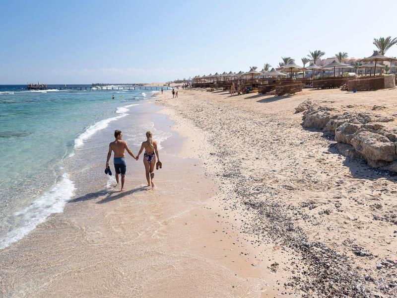Couple walking hand-in-hand on a sunny beach with clear water and palm trees nearby.