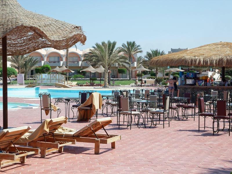 Outdoor pool area with loungers, straw umbrellas, bar seating, and palm trees in the background.