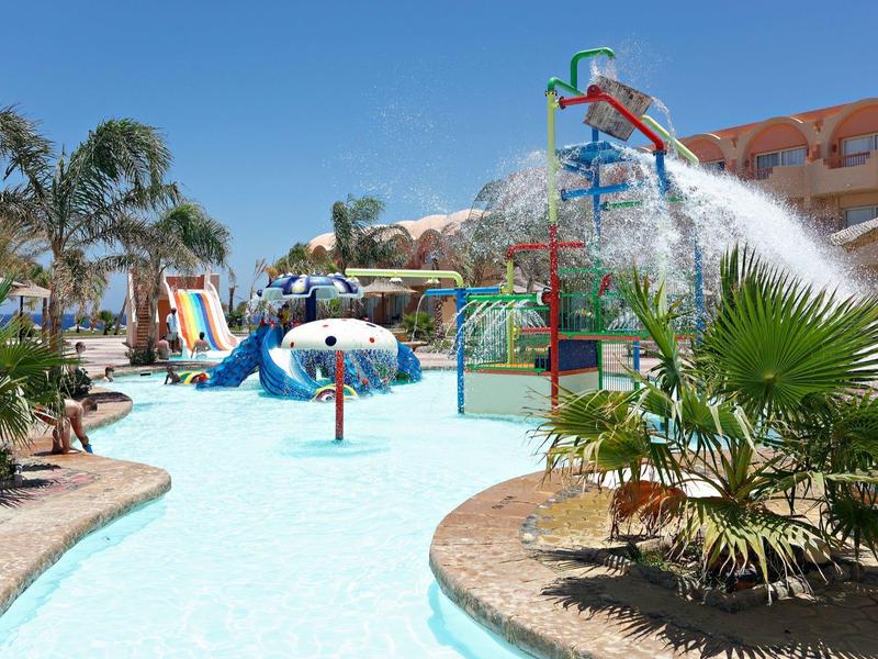 Outdoor water park with slides, fountains, and shallow pool surrounded by palm trees and buildings.