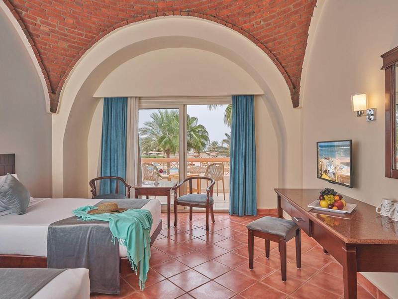 Hotel room with arched ceiling, two beds, desk, chairs, and balcony view of palm trees.