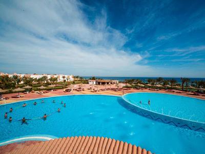 Gran piscina del hotel con hamacas azules bajo un cielo azul brillante junto al mar.