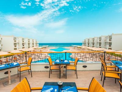 Terasa s stolky a židlemi s výhledem na hotelový bazén a moře pod modrou oblohou.