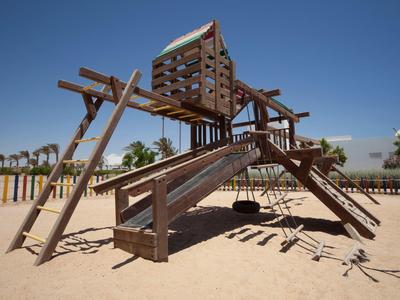 Estructura de juegos de madera en área de arena con columpios y toboganes bajo cielo azul.