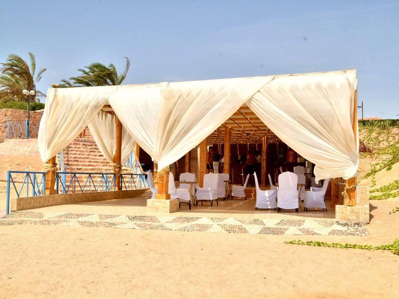 Pabellón abierto con cortinas blancas y sillas en la playa de arena bajo cielo azul.
