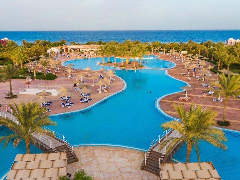 Gran piscina curva del hotel con tumbonas, palmeras y vista al mar al fondo.