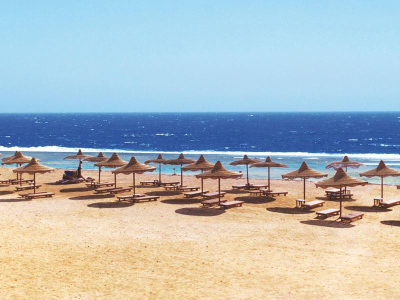 Strand mit vielen Sonnenschirmen aus Stroh, goldener Sand und blaues Meer im Hintergrund.