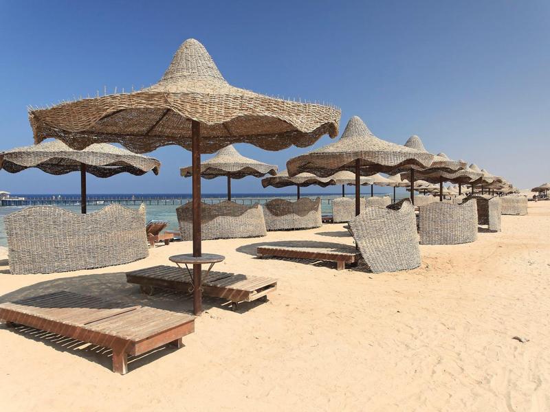 Sandstrand mit mehreren Strohmsonnenschirmen und Holzliegen unter klarem, blauem Himmel.