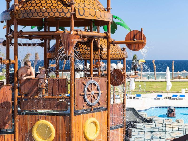 Kinder spielen auf einem hölzernen Piratenschiff neben einem Pool mit blauem Meer im Hintergrund.
